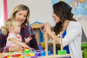 Caucasian mother and daughter work with preschool teacher. They are sitting at a table in the classroom working with educational toys.